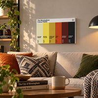 A framed 'The Birmingham Colour Palette' wall art print hangs on a beige wall above a sofa in a styled living room. The print displays six vertical color blocks: Old Bus Cream, Mr Egg Yellow, Jewellery Quarter Gold, Tikka Red, The Bull Bronze, and Sabbath Black, with CMYK codes and brief descriptions.